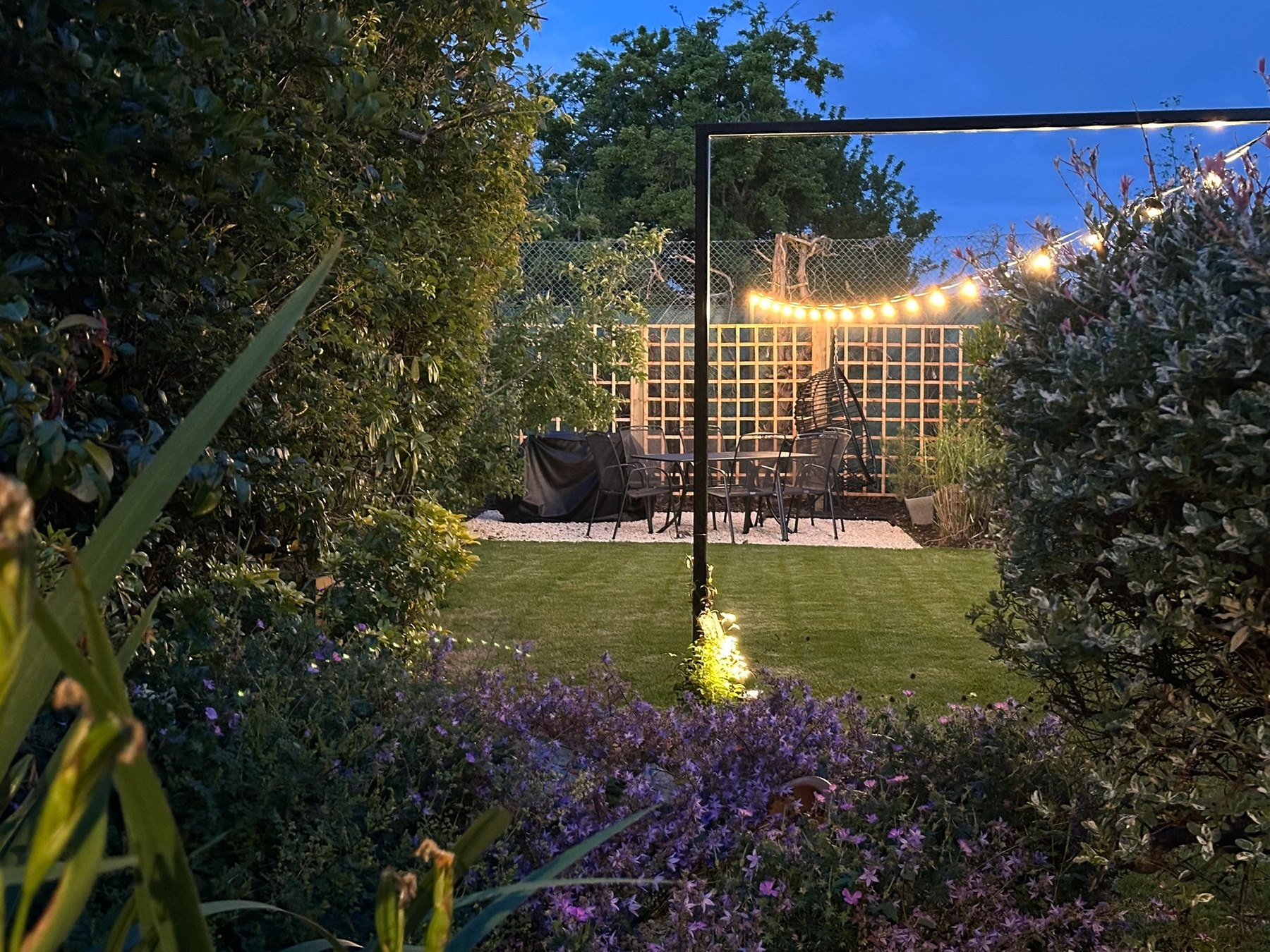 Layered garden planting, uplights and festoon lights around the black metal portal at dusk