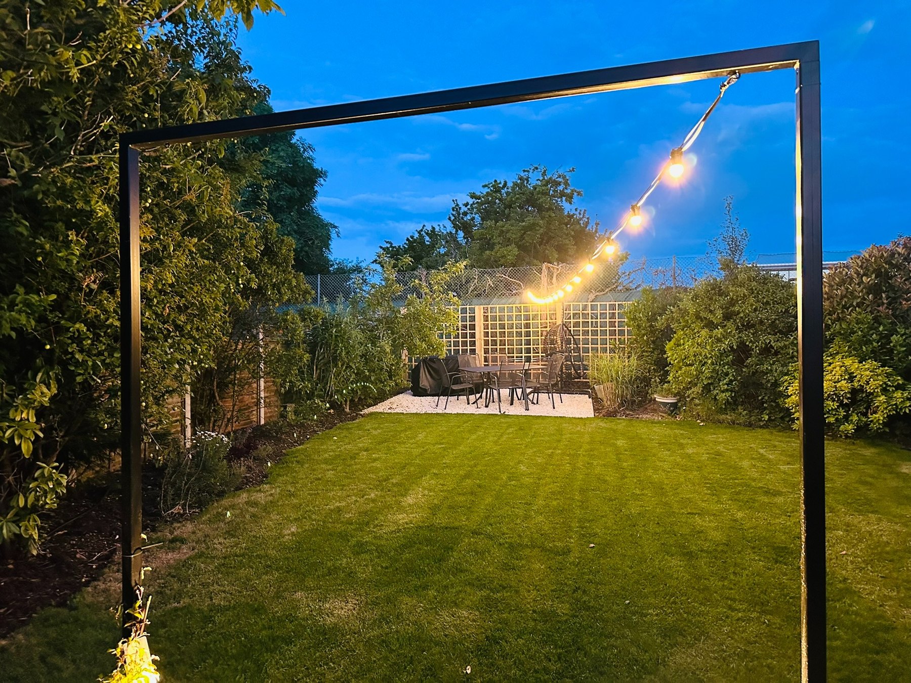 Mature garden at dusk with black metal portal, uplights and festoon lights leading to the outdoor dining area